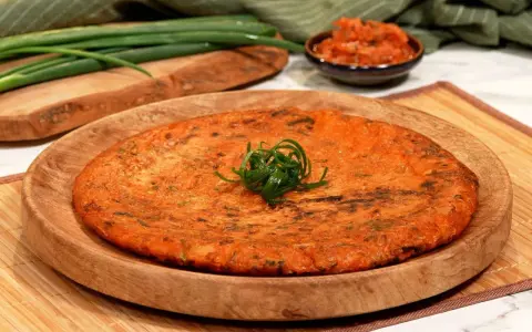 Ein goldbrauner, herzhafter Kimchi-Pfannkuchen, garniert mit Frühlingszwiebeln, liegt auf einem Holzteller. Daneben eine Schale mit Kimchi und grüne Frühlingszwiebeln.