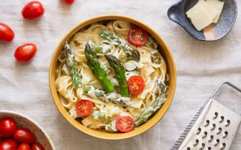 Pyszny makaron ze szparagami w kremowym sosie, ozdobiony pomidorkami koktajlowymi. Obok miseczka z parmezanem i tarką.