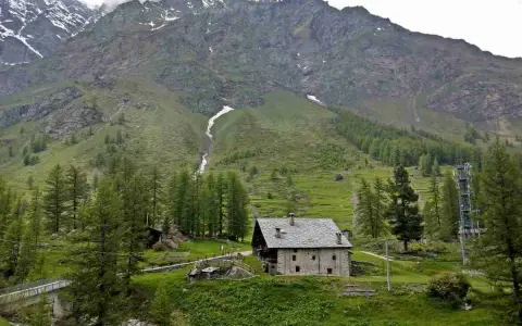 Un casolare in pietra nel **Val di Rhemes**, circondato da verdi pendii e imponenti montagne innevate.