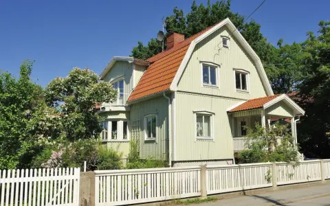 Ljusgrönt trähus med tegeltak och vit staket. En vacker trädgård med blommande buskar. Planerar att byta fasad för att fräscha upp huset.