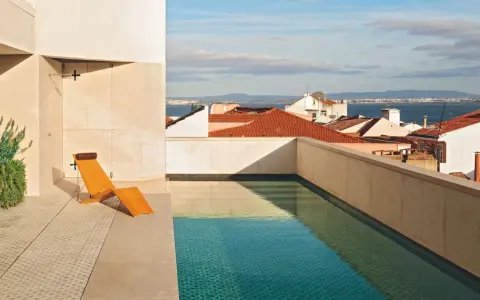 Una piscina en un balcón con vistas a tejados y al mar. Una tumbona de madera espera junto al agua cristalina.