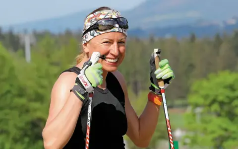 Uśmiechnięta Justyna Kowalczyk-Tekieli z kijkami do nartorolkarstwa, w sportowym stroju i opasce na głowie, gotowa do treningu.