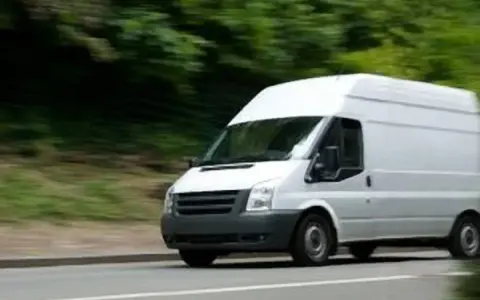 Furgone bianco in movimento su strada, con alberi sfocati sullo sfondo. Potrebbe essere un veicolo soggetto all'articolo 82 del codice della strada.