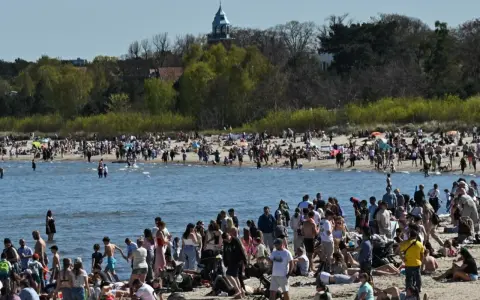 Tłumy ludzi na plaży, woda i zielone drzewa w tle. Widać wieżę kościelną. Ceny nad morzem pewnie wysokie, ale widok piękny.