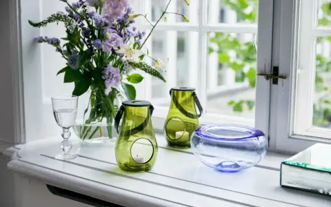 Ein Strauß lila Blumen und grüne Windlichter schmücken die Fensterbank. Ein Glas Wasser und eine blaue Schale runden die Dekoration ab.