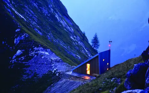 Un moderno bivacco interno, illuminato da una luce calda, si staglia su un pendio montuoso al crepuscolo.