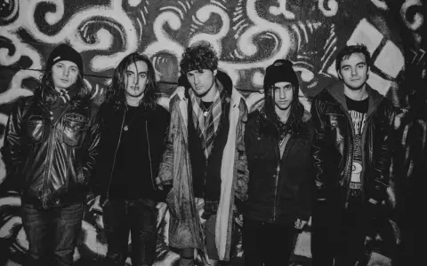 Fem unga män i rockkläder poserar framför en graffiti-vägg. De ser ut som ett band från 90 tals rock.