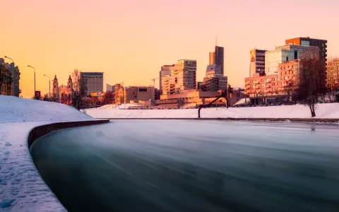 Zimowa rzeka w Wilnie o zachodzie słońca, z ośnieżonymi brzegami i nowoczesną panoramą miasta w tle.