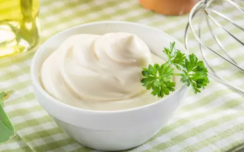 Un bol de mayonnaise maison onctueuse, garni de persil frais. Des œufs et un fouet à proximité suggèrent la préparation.