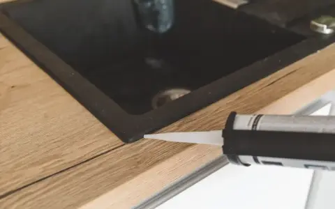 Pistolet z silikonem aplikuje uszczelnienie wokół zlewu, pokazując, jak polożyć silikon na blacie kuchennym.