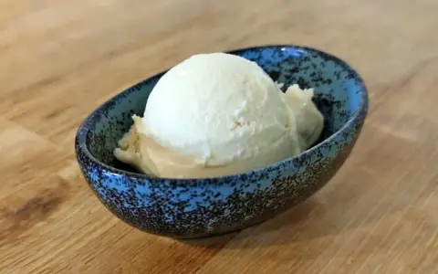 Ein Löffel cremiges Mandel Eis in einer blauen Schale auf Holztisch.