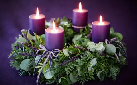 Four purple candles glow on a lush green Advent wreath, adorned with ribbons. The final candle's flame flickers, marking the 4th Advent.