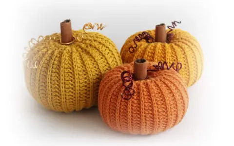 Tre virkade pumpor i höstfärger, en orange och två senapsgula, med kanelstänger som stjälkar och metalltråd som dekoration.