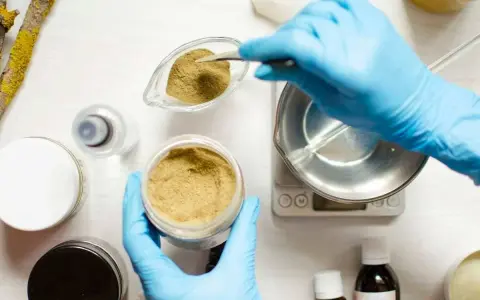 Ręce w niebieskich rękawiczkach odmierzają proszek do wagi, przygotowując składniki kosmetyczne, w tym butylene glycol.