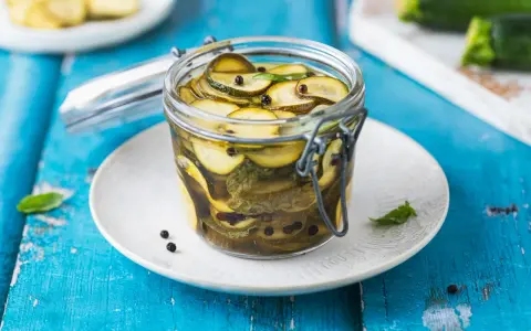 Barattolo di zucchine in agrodolce, con pepe nero e foglie di menta, su un piatto bianco appoggiato su un tavolo blu.