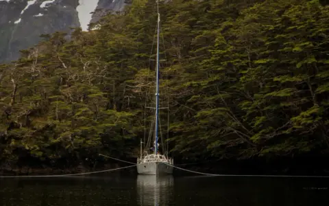 Voilier ancré dans un fjord, entouré d'une forêt dense. La chaîne du cablot, avec son épissure à 3 torons, assure la stabilité.
