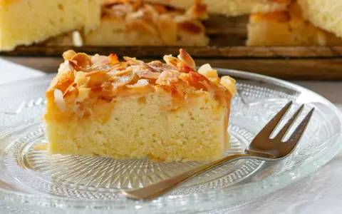 Ein Stück saftiger **Mandelkuchen vom Blech mit gemahlenen Mandeln** und Zucker bestreut, auf einem Glas-Teller mit einer Gabel.