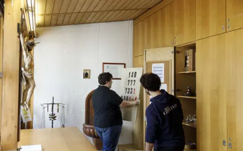 Zwei Männer im katholischen Priesterseminar hängen eine Liste mit Zahlen auf. Ein Kruzifix hängt an der Wand.