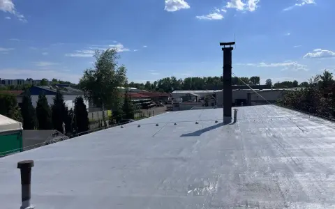 Nowy dach z doskonałą hydroizolacją, gotowy na każde warunki. W tle budynki przemysłowe i błękitne niebo z chmurami.