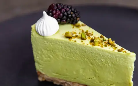 Pyszny sernik pistacjowy na zimno, udekorowany pianką, jeżyną i posypany pistacjami.