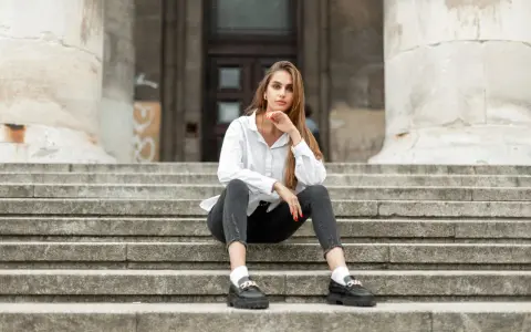 Eleganckie mokasyny to idealne buty do szarych spodni. Kobieta w białej koszuli i czarnych jeansach siedzi na schodach.