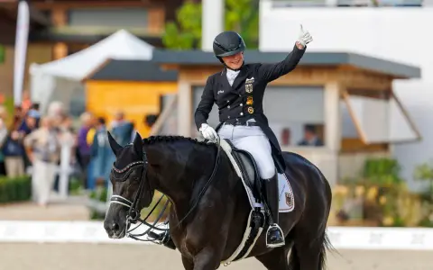 Reiterin gibt Daumen hoch nach erfolgreichem Grand Prix Reiten. Dunkles Pferd mit Turnieroutfit und Helm.