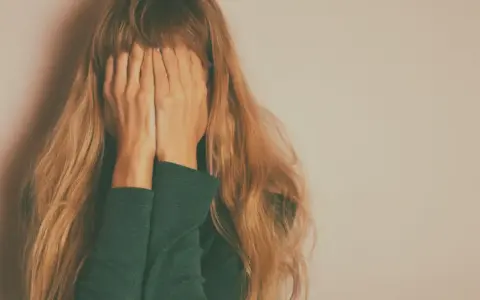 Kobieta z długimi blond włosami zakrywa twarz dłońmi na tle jasnej ściany.
