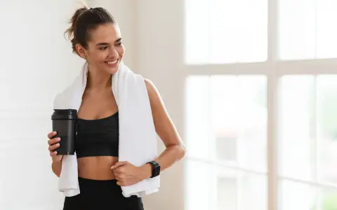 Lächelnde Frau mit Handtuch und Shaker nach dem Training. Sie fragt sich, wie viel Kreatin pro Tag für optimale Ergebnisse.