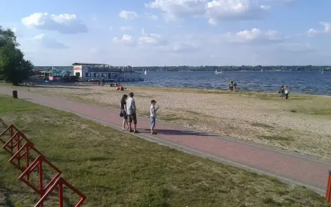 Rodzina spaceruje brzegiem jeziora Balewskiego. Na wodzie żaglówki, na brzegu barka z restauracją.