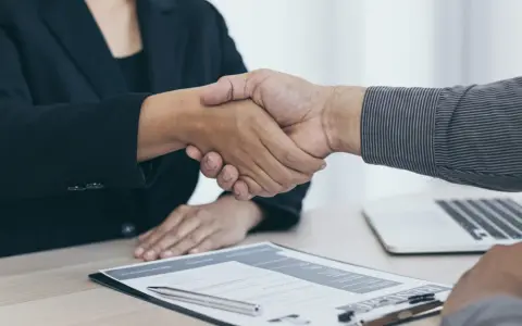 Dos personas se dan la mano sobre un escritorio con un currículum y un portátil. Es el final de una entrevista en francés.