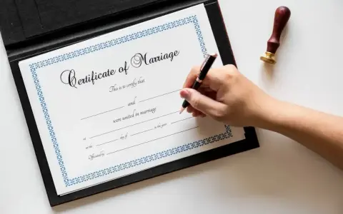 Dłoń pisze piórem na dokumencie "Certificate of Marriage", który poświadcza zawarcie związku małżeńskiego.