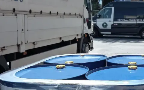 Niebieskie beczki z żółtymi zamknięciami, przygotowane do transportu towarów niebezpiecznych, obok ciężarówki i radiowozu Inspekcji Transportu Drogowego.