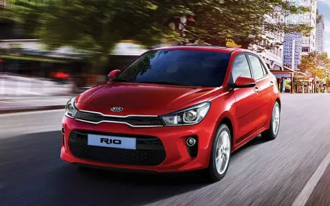 Un hatchback rojo Kia Rio se desplaza por una calle urbana, con edificios y árboles borrosos por el movimiento.