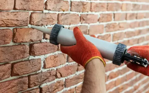Ręce w pomarańczowych rękawiczkach używają pistoletu do fugowania, by wypełnić ubytki w ceglanej ścianie.