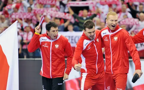 Trzech najlepszych piłkarzy ręcznych Polski w czerwonych strojach świętuje zwycięstwo. W tle kibice z biało-czerwonymi flagami.