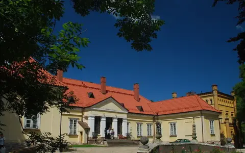 Pałac w Waplewie Wielkim, żółty, z czerwonym dachem, otoczony zielenią, pod błękitnym niebem.