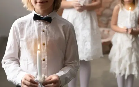 Dziecko w białej koszuli z czarną muszką trzyma zapaloną świecę. W tle inne dzieci w białych strojach.