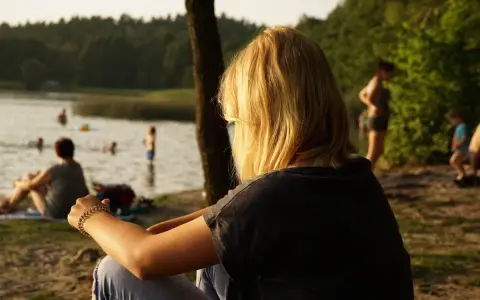 Dziewczyna z blond włosami siedzi nad jeziorem Trzechowskim, obserwując ludzi kąpiących się w wodzie.