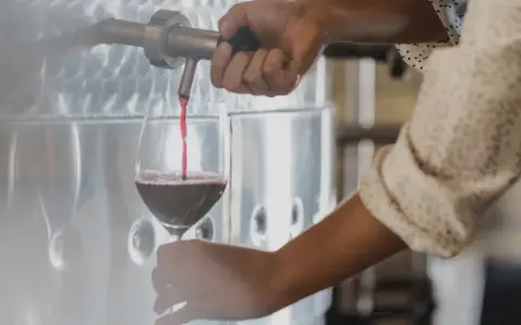 Rött vin hälls upp i ett glas från en stor tank. En hand vrider på kranen, kanske för att kontrollera tillsatser i vin.