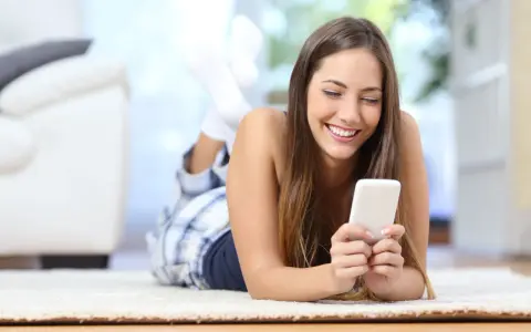 Uśmiechnięta dziewczyna w piżamie leży na dywanie, przeglądając telefon. Może szuka nowych znajomości na Tinder randki.