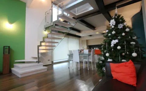 Un moderno soppalco casa con scala in vetro e legno, albero di Natale addobbato e un divano con cuscino arancione.