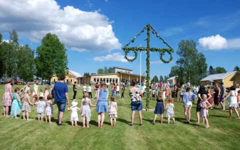 Många människor, både vuxna och barn, håller varandra i händerna och dansar runt en midsommarstång. De firar midsommar i en grönskande park med gula byggnader i bakgrunden.