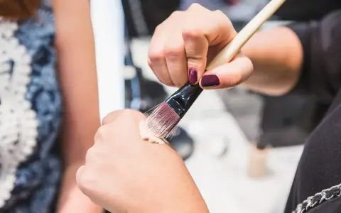 Dłoń z ciemnym lakierem do paznokci nabiera krem BB na pędzel, by nałożyć go na drugą dłoń.