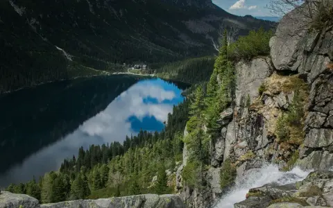 Widok z góry na Morskie Oko, odbijające chmury. Trasa turystyczna prowadzi przez skaliste zbocza z wodospadem.