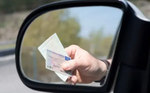 Mano sujetando un carnet de conducir y un documento de identidad reflejados en el espejo retrovisor de un coche.
