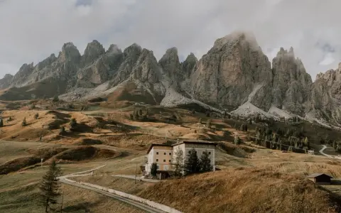 Malowniczy krajobraz w dolinie Val di Funes z budynkiem hotelowym na wzgórzu i skalistymi szczytami w tle.