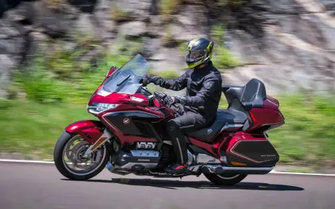 Mężczyzna w kasku jedzie lśniącym, czerwonym motocyklem uniwersalnym. W tle rozmyte zielone wzgórza i skały.