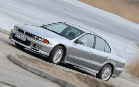 Srebrny Mitsubishi Galant z gracją pokonuje zakręt. Kierowca w okularach pewnie prowadzi auto.