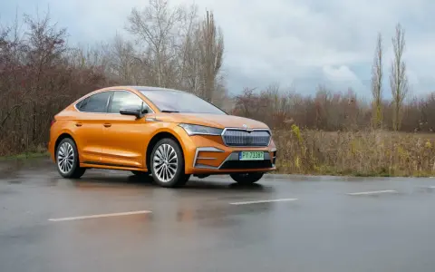 Pomarańczowa Skoda Enyaq Coupe na mokrej drodze, zarośla w tle.