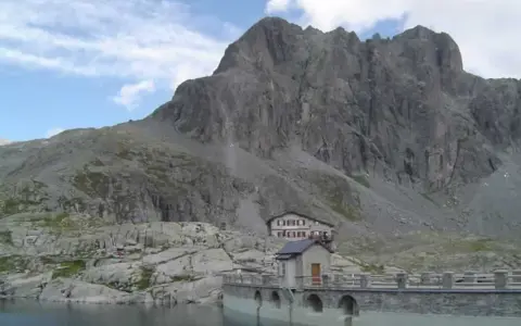 Rifugio sul lago della Vacca, con imponente parete rocciosa alle spalle.